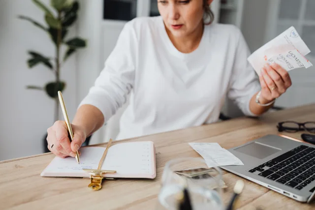 Woman managing finances, writing on notepad, and holding receipts at a desk. Portable restroom rental bookkeeping: cash flow basics